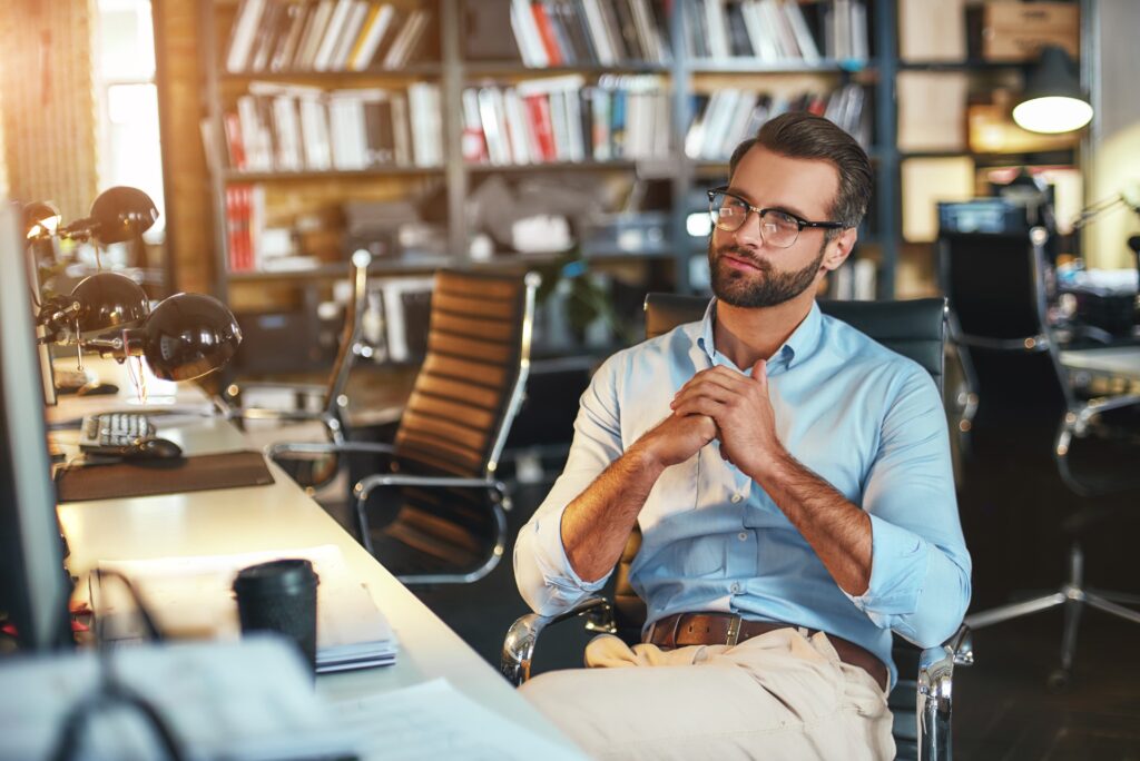 Lost in thoughts. Young bearded businessman in eyeglasses and formal wear working on computer and thinking about something while sitting in modern office. Work concept. Business concept