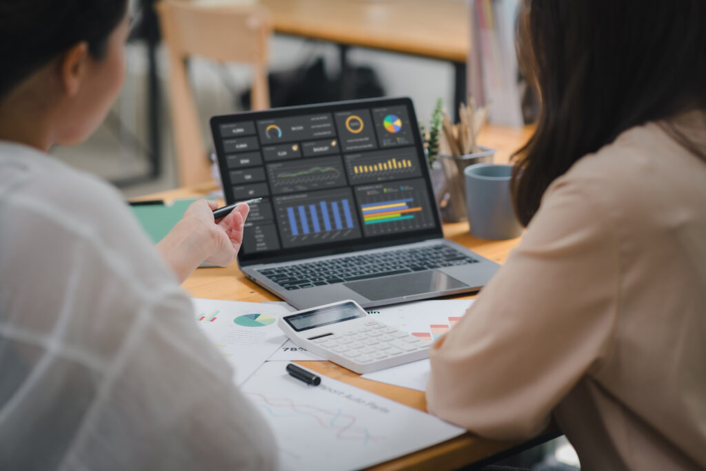 Two businesspeople or an accountant team are analyzing data charts, graphs, and a dashboard on a laptop screen in order to prepare a statistical report and discuss financial data in an office